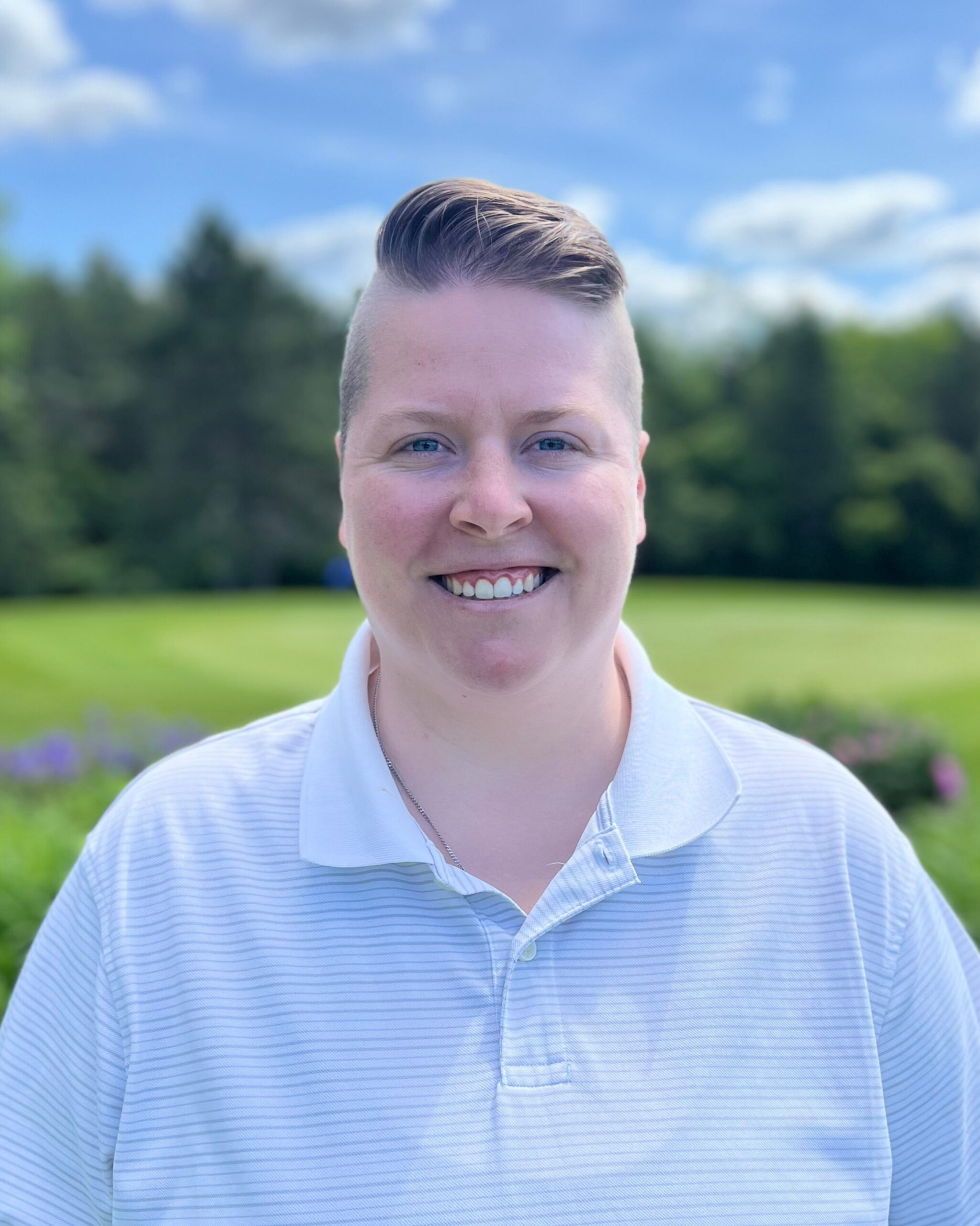 A person with short hair in a white polo shirt smiles warmly against a lush green landscape.